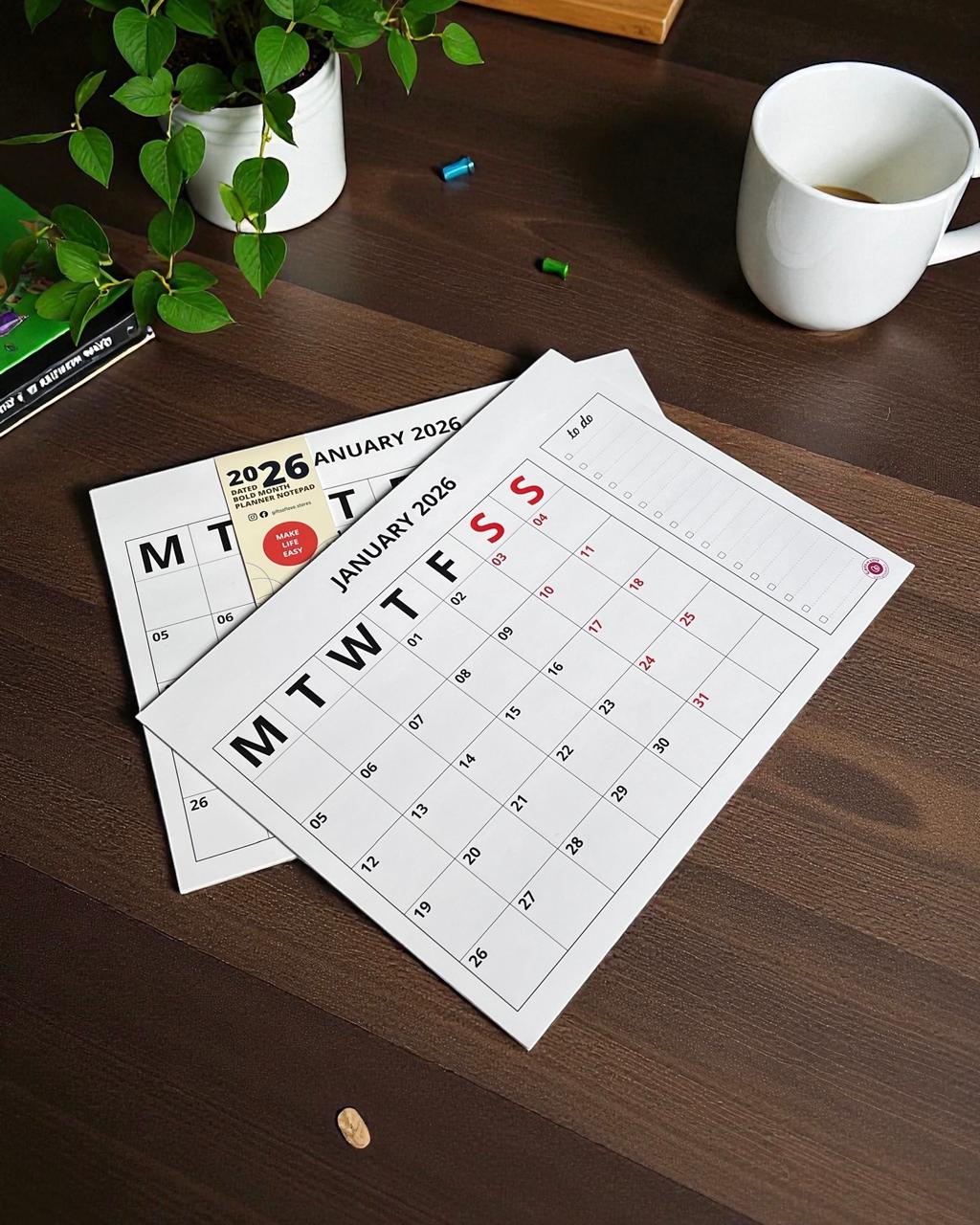 Calendars for January 2026 on a wooden surface with a cup and plant.