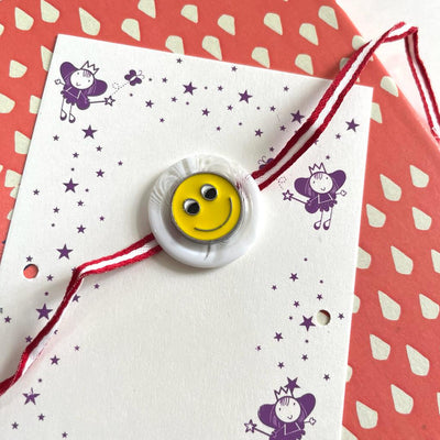 Gifts of Love Happy Little Fellow Kids Rakhi | Metal Smiley  on a red stripped ribbon.